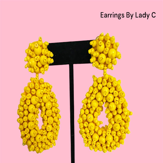 Yellow earrings on a black stand with a pink background, labeled 'Earrings By Lady C'.