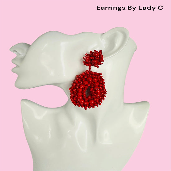Red beaded earrings on a white mannequin ear with a pink background, branded 'Earrings By Lady C'.