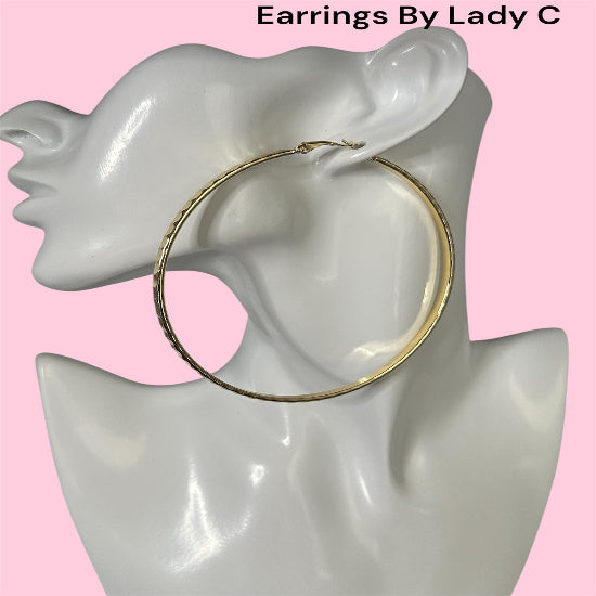 Gold hoop earring on a mannequin ear with 'Earrings By Lady C' text above, against a pink background